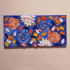 May include: A rectangular canvas painting featuring a vibrant floral design. The artwork showcases textured, raised flowers in shades of white, orange, and blue, set against a deep blue background. Scattered petals in various colors add depth.