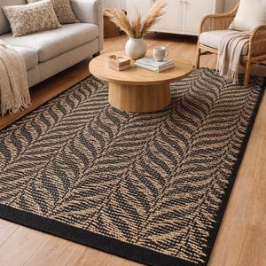 Könnte beinhalten: Rechteckiger Teppich mit schwarzem Rand und sich wiederholendem Blattmuster in Schwarz und Beige. Der Teppich liegt auf einem Holzboden, und die Szene umfasst ein Sofa, einen Couchtisch und einen Korbstuhl.