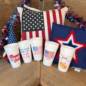 May include: Five white plastic cups with patriotic designs. One says Happy 4th of July, one says You Look Like The Fourth of July, one features an American flag with The Martins 2026, and one says Bad Day To Be A Hot Dog with a hot dog graphic.