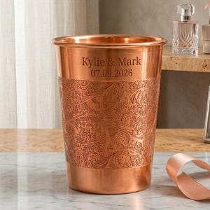 Puede incluir: Un vaso de metal color cobre con un intrincado patrón floral en relieve alrededor de la parte inferior y el texto grabado "Kylie & Mark" seguido de la fecha "07.09.2026" cerca del borde superior del recipiente.