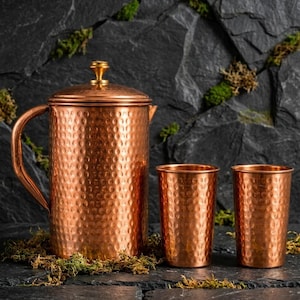 Puede incluir: Una jarra de cobre martillado con tapa y asa a juego, acompañada de dos vasos de cobre martillado. El conjunto presenta un acabado metálico uniforme con un patrón de superficie texturizado y con hoyuelos en las tres piezas.