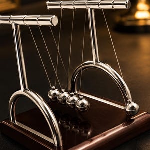 Peut inclure: Un pendule de Newton composé de cinq sphères métalliques argentées polies, suspendues par de fins fils à deux cadres arqués argentés. L'ensemble est fixé sur une base rectangulaire en bois brun foncé avec une finition lisse et réfléchissante.