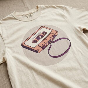 May include: Cream-colored t-shirt featuring a retro cassette tape design. The tape is peach and brown, with the text "for rainy days only" printed on it. The tape is set against a light purple circle.