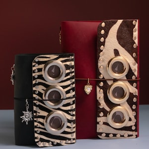 Peut inclure: Deux carnets rectangulaires avec des couvertures en cuir. Le plus petit est noir et le plus grand est rouge foncé. Ils présentent une bande verticale de fourrure à motif zèbre, trois grands œillets métalliques argentés et des clous argentés.