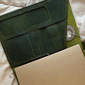 May include: A dark green leather journal cover with multiple interior pockets and slots, featuring a light brown paper notebook insert. The cover is accented with silver-toned metal studs, a circular charm, and a silver-toned wing-shaped pendant.