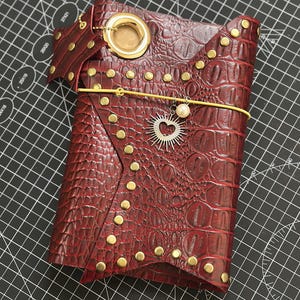Op de afbeelding: Een donkerrode notitieboekhoes met krokodillenleertextuur, voorzien van goudkleurige metalen studs langs de randen, een grote gouden oogje aan de bovenkant en een gele elastische bandsluiting. Een zilveren hartvormige zonhanger en een witte parel zijn bevestigd.