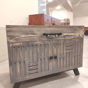 Puede incluir: Un mueble de madera gris con un diseño geométrico en las puertas. El mueble tiene un tirador negro y patas negras. La veta de la madera es visible y el mueble parece hecho a mano.