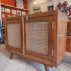 Puede incluir: Un mueble de madera con dos puertas y paneles de mimbre tejido. El mueble tiene un acabado de madera natural y patas anguladas. Las puertas tienen tiradores verticales de madera y un cierre de pestillo.