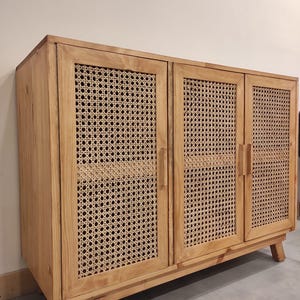 Puede incluir: Un mueble de madera clara con tres puertas. Cada puerta presenta un panel de ratán tejido enmarcado por la madera. El mueble se apoya en patas de madera anguladas, ofreciendo una estética moderna de mediados de siglo.