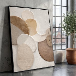 Puede incluir: Impresión artística abstracta con una superficie texturizada y círculos superpuestos en tonos beige, marrón y blanco. La obra de arte está enmarcada en un marco negro y se apoya en una pared gris. La obra está cerca de una ventana con vistas a una ciudad.