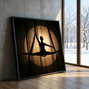May include: Framed art print featuring a silhouette of a person performing aerial silks. The figure is centered in a spotlight, with the background showing a wooden texture. The print is in a black frame, leaning against a concrete wall.