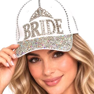 May include: A white baseball cap featuring the word BRIDE in large silver-toned rhinestones, topped with a rhinestone tiara graphic. The brim is covered in multi-colored iridescent glitter, and the seams are lined with small silver studs.