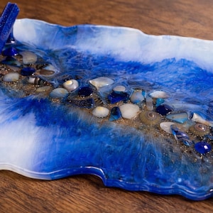 May include: A rectangular resin tray with a blue and white ocean-like design. The tray is embedded with seashells, blue stones, and gold glitter. A blue handle is attached to the side. The tray is approximately 12 inches long.