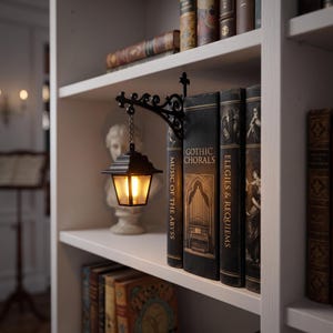May include: A black metal wall-mounted lantern with a chain, emitting a warm glow, attached to a book titled Gothic Chorals. The lantern features a square frame with frosted glass panels and is positioned on a white shelf among other books.