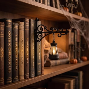 May include: A black metal wall-mounted bracket with ornate scrollwork, featuring a small hanging lantern with a warm glowing light. The bracket is positioned between several vintage-style books with titles like Gothic Chronicles and Organ Worlds.