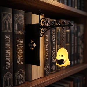 May include: A black metal wall-mount bracket with a decorative scroll design holds a small, glowing yellow ghost figure suspended by a chain. The ghost is shaped like a rounded character holding a tiny black book, positioned between two books.
