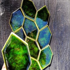 Könnte beinhalten: Handgefertigte, sechseckige Keramikfliesen mit glänzendem, irisierendem Finish. Die Fliesen zeigen eine Mischung aus Grün- und Blautönen, die einen einzigartigen, organischen Look erzeugt. Die Fliesen sind auf einer verwitterten Holzoberfläche angeordnet.