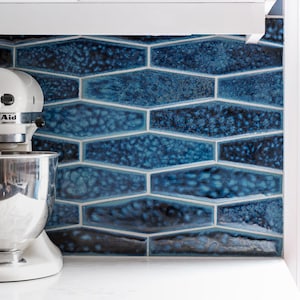Könnte beinhalten: Ein weißer KitchenAid-Küchenmixer mit einer Edelstahlschüssel. Im Hintergrund befindet sich eine blaue, geometrische Fliesenrückwand. Die Fliesen haben ein strukturiertes, gesprenkeltes Aussehen. Der Mixer steht auf einer weißen Arbeitsplatte.