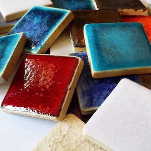 Può includere: Una collezione di piastrelle quadrate in ceramica in vari colori, tra cui tonalità di blu, rosso, marrone e bianco. Le piastrelle hanno una superficie lucida e strutturata, disposte in una ripresa ravvicinata.