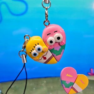 May include: Heart-shaped keychains featuring SpongeBob and Patrick Star characters, split into two halves. The keychains are yellow and pink with cartoon details. A smaller heart-shaped keychain is also present, split into two halves with the names "Alex" and "Max".