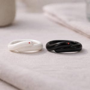 May include: Two rings in the shape of snakes, one white and one black, each with a small red dot. The rings are displayed on a light-coloured, textured surface. The rings are likely made of ceramic or a similar material.