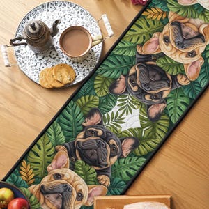May include: A decorative table runner featuring a repeating pattern of French bulldogs amidst lush green tropical leaves. The runner has a black border and is placed on a wooden surface. A tray with a cup of coffee, cookies, and a teapot is visible.