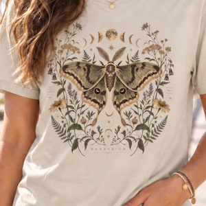 Peut inclure: Un t-shirt beige à manches courtes orné d'un motif central représentant un grand papillon de nuit entouré d'illustrations botaniques de fleurs, de fougères et d'un cycle lunaire. Le mot "HERBARIUM" est inscrit en bas du motif.