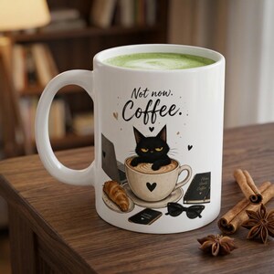 Könnte beinhalten: Ein weißer Keramikbecher gefüllt mit grünem Matcha-Tee, verziert mit einer Grafik einer mürrischen schwarzen Katze in einer Kaffeetasse. Der Text lautet Not now. Coffee. Zu sehen sind zudem ein Laptop, ein Croissant, ein Notizbuch und eine Sonnenbrille.