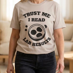 Könnte beinhalten: Beiges T-Shirt mit dem schwarzen Text "TRUST ME I READ LAB RESULTS". Das Design beinhaltet ein rundes Grafik mit drei dunklen Formen. Das kurzärmlige T-Shirt wird mit dunklen Jeans getragen.