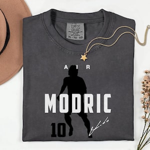 a gray t-shirt with a silhouette of a person and the text "MODRIC 10" printed on it. The t-shirt is laid out on a white surface, accompanied by a hat, a necklace, and some flowers.