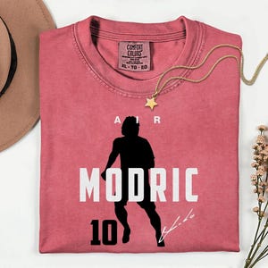 a pink t-shirt with a silhouette of a person and the text "MODRIC 10" printed on it.