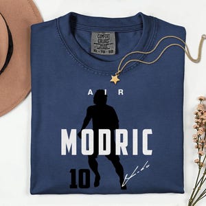 A blue t-shirt with a silhouette of a person and the text "MODRIC 10" printed on it, along with a gold star necklace and a brown hat.