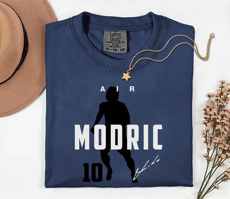A blue t-shirt with a silhouette of a person and the text "MODRIC 10" printed on it, along with a gold star necklace and a brown hat.