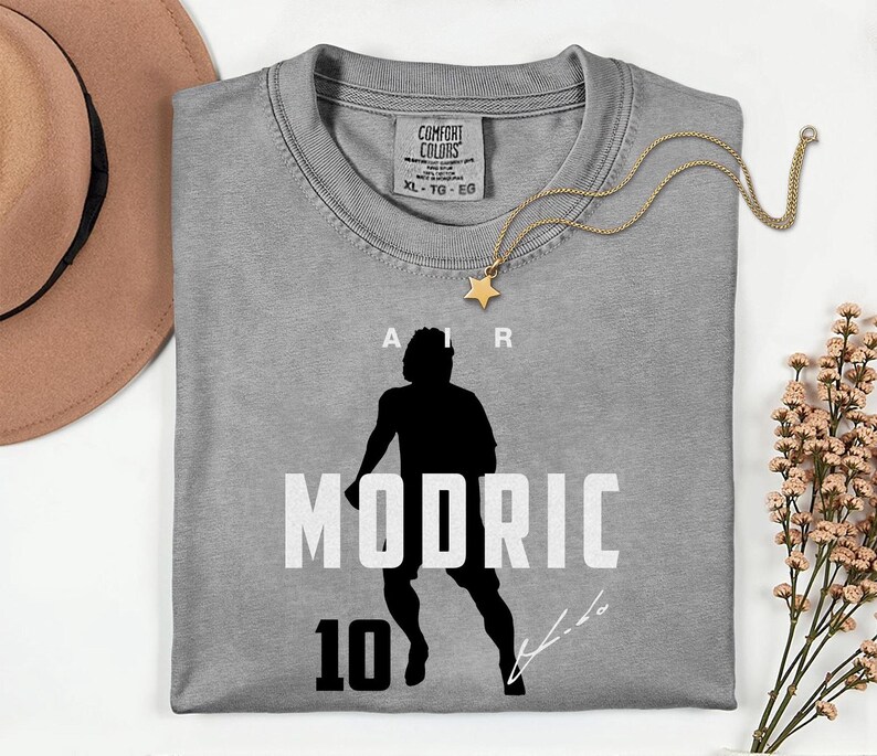 a gray t-shirt with a silhouette of a person and the text "MODRIC 10" printed on it. The t-shirt is laid out on a white surface, accompanied by a hat, a necklace, and some flowers.