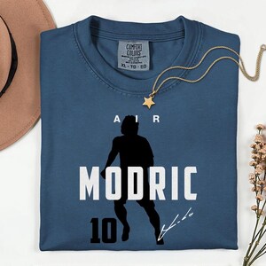 A blue t-shirt with a silhouette of a person and the text "MODRIC 10" printed on it, along with a gold star necklace and a brown hat.