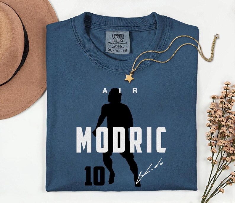 A blue t-shirt with a silhouette of a person and the text "MODRIC 10" printed on it, along with a gold star necklace and a brown hat.