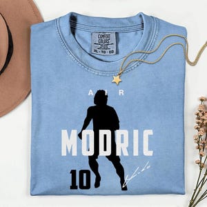 A blue t-shirt with a silhouette of a person and the text "MODRIC 10" printed on it, along with a gold star pendant.