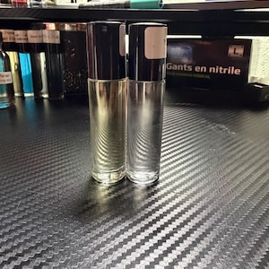May include: Two cylindrical glass roller bottles with black caps standing upright. The left bottle contains a pale yellow liquid, while the right bottle is empty and clear. Each cap features a small white rectangular label on the side.
