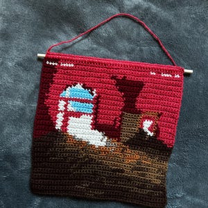 May include: A rectangular crochet wall hanging featuring a pixelated landscape design. The scene displays a red sky, a brown ground, and a central structure with white and blue details. It is suspended from a wooden dowel by a red yarn string.