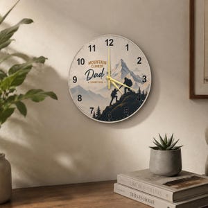 Op de afbeelding: Een ronde wandklok met een thema van bergbeklimmen. De wijzerplaat toont een illustratie van een bergketen met de woorden "MOUNTAIN CLIMBER Dad SUMMIT RING". De wijzers van de klok zijn geel.