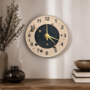 May include: A round wall clock with a cream-colored face and gold hands. The clock features a solar system design with planets and a dark blue center. The numbers are in black, and the clock is mounted on a light-colored wall.