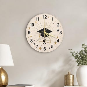 May include: A round wall clock with a cream-colored face. The clock features a black graduation cap, a scroll, and gold star accents. The clock hands and numbers are gold. The clock is a decorative item.