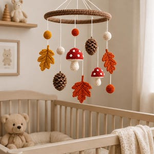 May include: A crochet nursery mobile featuring a brown circular frame suspended by white strings. Hanging elements include two red and white mushrooms, two brown pinecones, three autumn leaves in orange and yellow, and several small spheres.