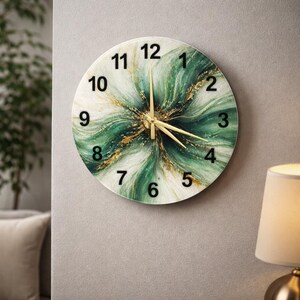 Puede incluir: Reloj de pared redondo con un diseño floral abstracto verde y dorado. La esfera del reloj presenta números negros y manecillas doradas. El reloj está montado en una pared gris.