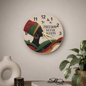 Puede incluir: Un reloj de pared redondo con la ilustración de perfil de una persona con un pañuelo rojo, verde y amarillo y pendientes de aro. La esfera muestra el texto FREEDOM NEVER SLEEPS JUNETEENTH, números negros, manecillas doradas y aves.