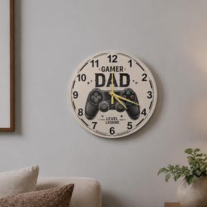Könnte beinhalten: Eine runde Wanduhr mit cremefarbenem Zifferblatt. Die Uhr zeigt die Worte "GAMER DAD" über einer schwarzen Gamecontroller-Grafik. Der Text "LEVEL LEGEND" befindet sich darunter.