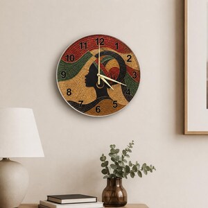 Puede incluir: Un reloj de pared redondo con una ilustración estilo mosaico de una persona de perfil que lleva un pañuelo en la cabeza. El diseño utiliza colores rojo, verde, amarillo y negro. Tiene números negros del 1 al 12 y manecillas doradas finas.