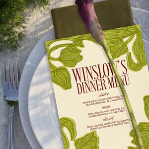 May include: A dinner menu titled "Winslow's Dinner Menu" with a green floral design, a purple calla lily, and a white plate. The menu lists a starter, main course, and dessert. The menu is on a white tablecloth.