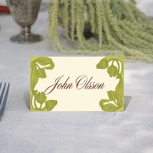 May include: A cream-colored place card with the name "John Olsson" in burgundy script. The card is decorated with green floral corner designs. The card is set on a white tablecloth, with a fork visible on the left.