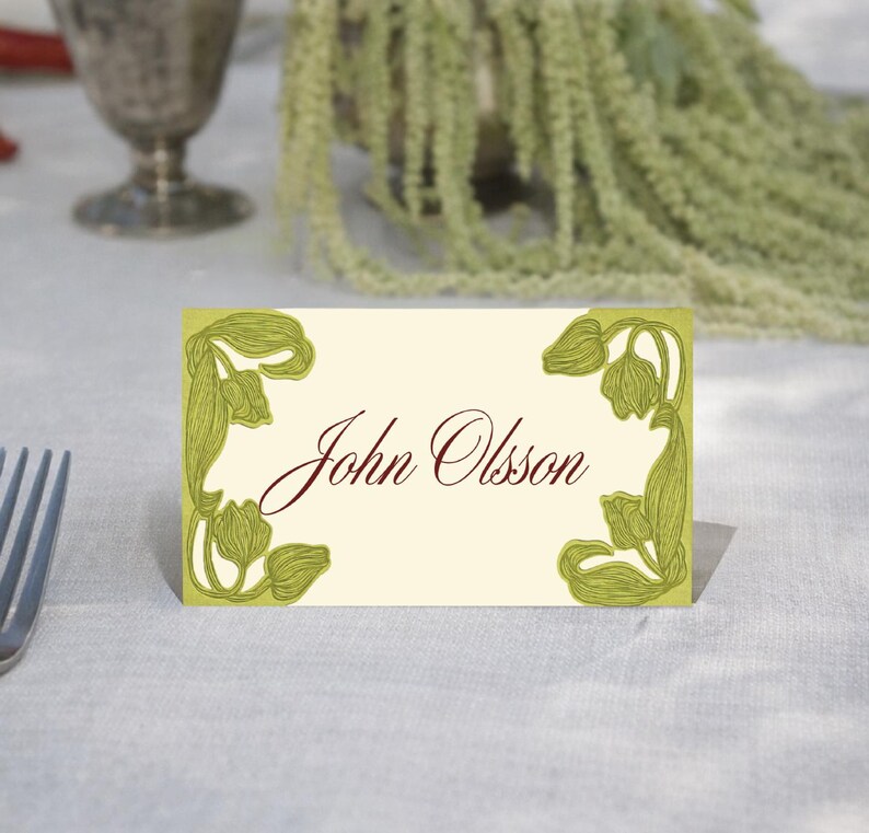 May include: A cream-colored place card with the name "John Olsson" in burgundy script. The card is decorated with green floral corner designs. The card is set on a white tablecloth, with a fork visible on the left.